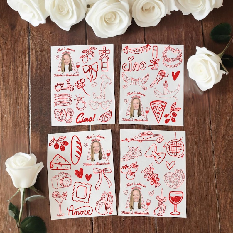 May include: Four white cards with red illustrations and text, including a bride's face, hearts, and Italian-themed drawings. The cards are arranged on a wooden surface, surrounded by white roses.