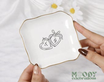 May include: A square white ceramic trinket dish with wavy edges and a gold-colored rim. The center features a black line art illustration of two connected heart-shaped lockets tied with a bow, with the letters T and G printed inside each heart.