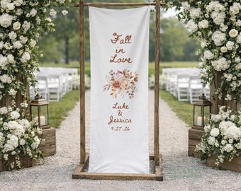 Puede incluir: Un estandarte de boda vertical de tela blanca suspendido de un marco de madera, con el texto Fall in Love, los nombres Luke & Jessica y la fecha 27.09.26 en letra color óxido, acentuado por una ilustración floral central en tonos otoñales.