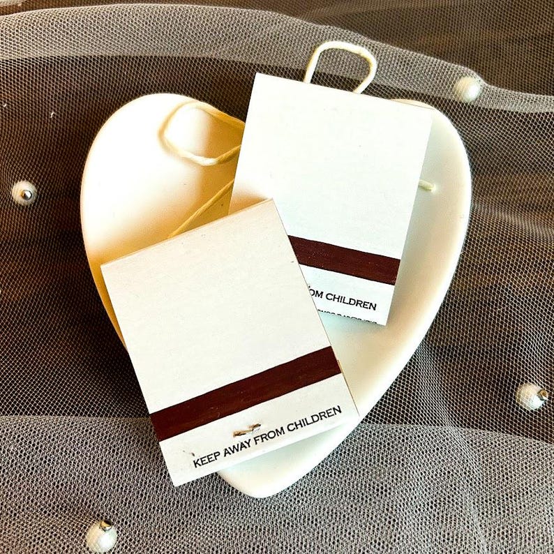 Puede incluir: Dos etiquetas rectangulares blancas con una franja marr&oacute;n oscuro y el texto "KEEP AWAY FROM CHILDREN" se colocan en un plato blanco en forma de coraz&oacute;n. Las etiquetas tienen un peque&ntilde;o lazo en la parte superior. El plato est&aacute; sobre una superficie transparente de color marr&oacute;n claro.