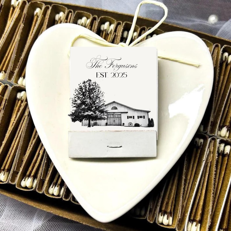 Puede incluir: Un plato de cer&aacute;mica en forma de coraz&oacute;n de color crema contiene una caja de cerillas con una ilustraci&oacute;n en blanco y negro de un &aacute;rbol y un granero. La caja de cerillas dice "The Fergusons EST. 2025". Las cerillas est&aacute;n en una caja de cart&oacute;n marr&oacute;n.