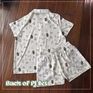 May include: White pajama set with a short-sleeved button-up shirt and matching shorts. The set is covered in a repeating pattern of black line drawings and brown faces. The text "Back of PJ Set" is at the bottom.