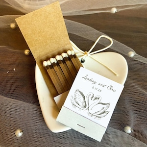 May include: A matchbox with brown matches and a white ceramic heart-shaped dish. The matchbox has the text "Lindsay and Chris 6.17.26" and a drawing of two swans. The background is a sheer, gray fabric with scattered pearls.