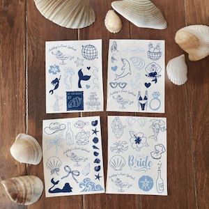 May include: Four sheets of blue and white temporary tattoos with beach-themed illustrations, including mermaids, seashells, and cocktails. The text "Ashley's Last Splash" and "Bride" are visible. Seashells surround the sheets on a wooden surface.