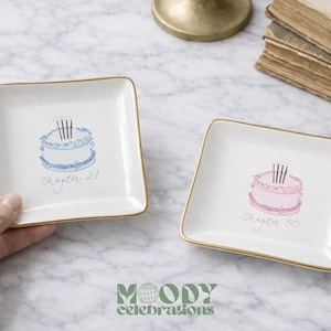 May include: Two square white ceramic trinket dishes with gold-toned rims. One dish features a blue cake illustration with the text Chapter 21, and the other features a pink cake illustration with the text Chapter 50, both with four candles.
