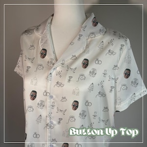 May include: White button-up top with a pattern of black line art wedding-themed illustrations and faces. The illustrations include wedding cakes, bells, rings, and doves. The text "Button Up Top" is at the bottom of the image.