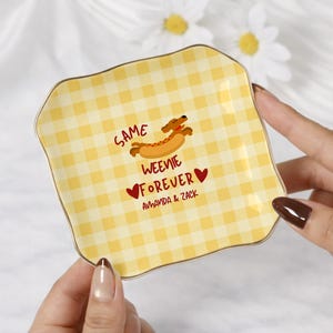 May include: A square ceramic trinket dish with a yellow and white gingham pattern, featuring a cartoon dachshund in a hot dog bun, red hearts, and the text SAME WEENIE FOREVER AMANDA & ZACK printed in the center with a gold-toned rim.