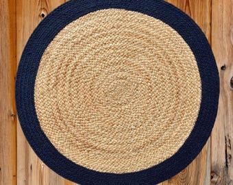 May include: A round jute rug with a navy blue border. The center of the rug is a natural tan color, with a spiral pattern. The border is a solid navy blue. The rug is placed on a wooden surface.
