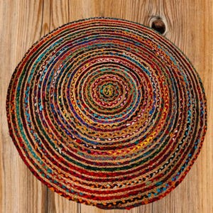 May include: A round, braided jute rug with a spiral design. The rug features a vibrant mix of colors, including red, blue, yellow, and green, creating a visually appealing pattern. The rug is placed on a wooden surface.
