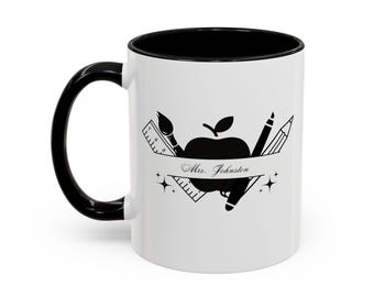 May include: White ceramic mug with a black handle and interior. The mug features a black graphic of an apple, pencil, paintbrush, and ruler with the text "Mrs. Johnston" on a banner. The design is surrounded by star shapes.