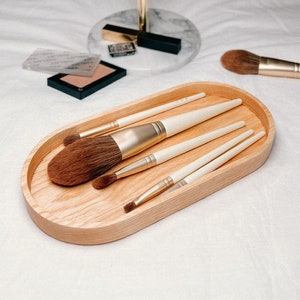 A wooden oval tray holding makeup brushes is on a bed, with a round gold-framed mirror and makeup items in the background.