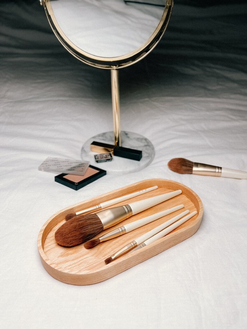 A wooden oval tray holding makeup brushes is on a bed, with a round gold-framed mirror and makeup items in the background.