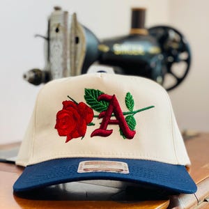 May include: A cream-colored baseball cap with a dark blue brim. The front features an embroidered design of a red rose with green leaves and a large, stylized red letter A. A rectangular sticker is attached to the center of the dark blue brim.
