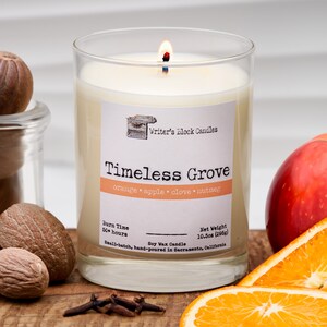 May include: A glass jar candle with a white label that reads "Writer's Block Candles Timeless Grove orange apple clove nutmeg Burn Time 50+ hours Soy Wax Candle Net Weight 10.5oz (298g) Small-batch, hand-poured in Sacramento, California".
