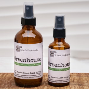 May include: Two amber glass bottles with black spray tops. Both bottles have a white label with the text "Writer's Block Candles Greenhouse carrot tomato thyme basil Room & Linen Spray Net Weight 4oz (113g) Small-batch, hand-poured in Sacramento, California".