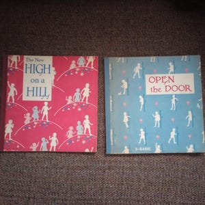 May include: Two vintage children's books. One book has a pink cover with the title "The New High on a Hill" and illustrations of children. The other book has a blue cover with the title "Open the Door" and illustrations of children.