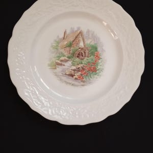 May include: White ceramic plate with a scalloped edge and embossed floral design. The center features a scenic illustration of a water mill with a stone exterior, surrounded by greenery and red flowers.