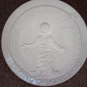 May include: A white ceramic plate with a raised relief design. The design features a child figure with a halo, arms outstretched, within a cross. The words "KING OF KINGS - 70" are inscribed around the edge. The plate is round.