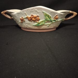 May include: A cream-colored ceramic serving dish with two handles, featuring a textured surface and a raised relief design of orange berries and green leaves. The base is a soft pink, and the handles are accented with brown tones.