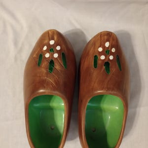 May include: A pair of brown wooden-style clogs featuring a bright green interior. The exterior of each clog is decorated with carved green accents and a floral pattern composed of five small white painted dots on the top surface.