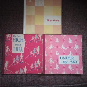May include: Three vintage children's books. The top book has a checkered pattern and the text "Skip Along". The other two books are pink with illustrations of children and the titles "The New High on a Hill" and "Under the Sky".