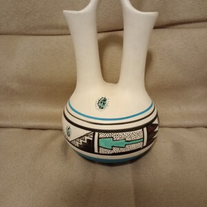 May include: A white ceramic vase with two openings, decorated with geometric patterns in black, turquoise, and red. The vase has a traditional Southwestern design.