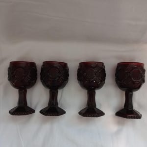 May include: Four ruby red goblets with ornate floral designs. Each glass features a detailed pattern on the bowl and a stemmed base. The glasses are arranged in a row against a white background, showcasing their vintage aesthetic.
