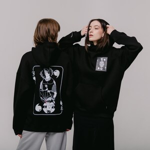 May include: Two black hoodies with a white graphic print of a playing card. The card features a person holding a staff and a symbol with a circle and a cross. The hoodie on the left has the card on the back and the hoodie on the right has the card on the front.