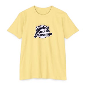 May include: A pale yellow short-sleeve t-shirt featuring a central circular graphic with a dotted background. The text Living Lavish Massage is printed in a dark blue, bold, cursive font across the center of the circle on the front of the shirt.