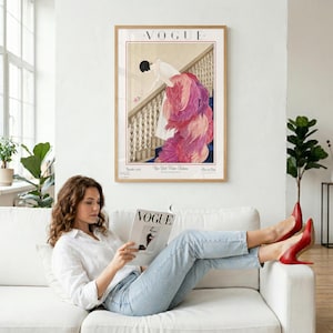 May include: A framed Vogue magazine cover art print. The artwork features a woman in a long, pink feathered gown descending a staircase. The print is in a wooden frame and hangs above a woman reading a Vogue magazine on a white sofa.