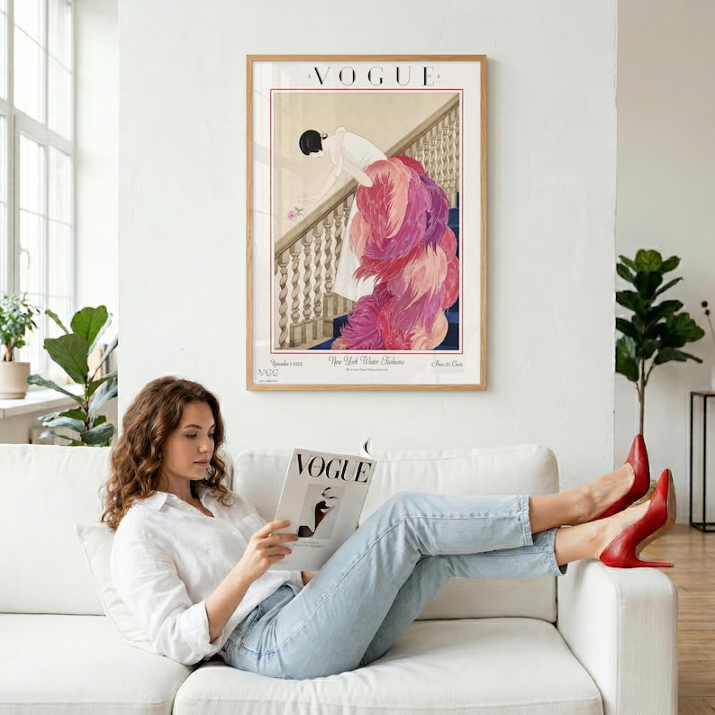 May include: A framed Vogue magazine cover art print. The artwork features a woman in a long, pink feathered gown descending a staircase. The print is in a wooden frame and hangs above a woman reading a Vogue magazine on a white sofa.