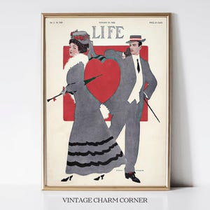 May include: A vintage LIFE magazine cover from February 20, 1908, featuring a couple in a romantic pose. The woman is wearing a gray dress with black trim and a hat with a red flower. The man is wearing a gray suit with a white shirt and a black tie. The cover features a large red heart in the background.