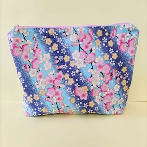 Può includere: Un beauty case rettangolare con motivo floreale. La borsa presenta uno sfondo blu con fiori di ciliegio rosa e bianchi e dettagli dorati. Ha una cerniera rosa e un interno azzurro.