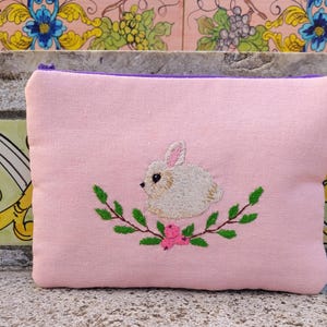 Può includere: Un astuccio rettangolare rosa con cerniera viola. L'astuccio presenta un disegno ricamato di un coniglietto bianco circondato da foglie verdi e fiori rosa. L'astuccio è realizzato in un tessuto morbido e strutturato.