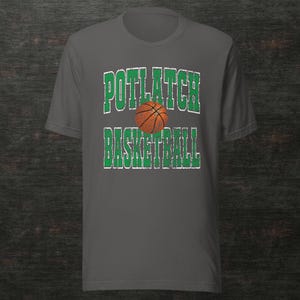May include: A grey t-shirt with the text "POTLATCH BASKETBALL" in distressed green lettering and a basketball graphic. The shirt has a crew neck and short sleeves. The design is centered on the front.