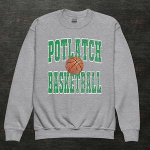 May include: A grey sweatshirt featuring the text "POTLATCH BASKETBALL" in green, distressed lettering, with a basketball graphic in the centre. The sweatshirt has a crew neck.