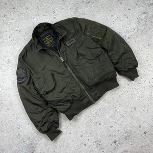 May include: An olive green bomber jacket featuring a full-length front zip, ribbed cuffs and hem, and two large front flap pockets. The jacket includes a utility pocket on the left sleeve, a small rectangular brand patch, and a circular patch.