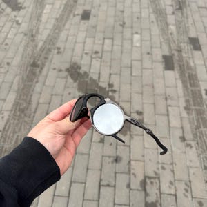 May include: Black and silver sunglasses with a unique circular lens design. The sunglasses have a sleek, modern frame and are held up against a gray brick background. The sunglasses are a stylish accessory.