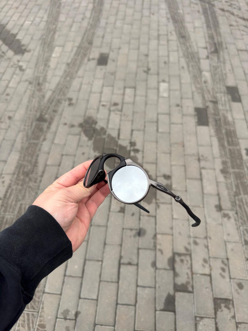 May include: Black and silver sunglasses with a unique circular lens design. The sunglasses have a sleek, modern frame and are held up against a gray brick background. The sunglasses are a stylish accessory.