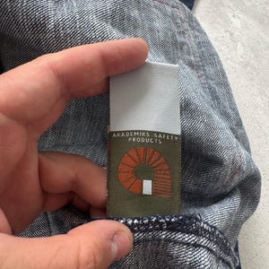 Może przedstawiać: Zbliżenie na tkaninę dżinsową z metką z napisem "AKADEMIKS SAFETY PRODUCTS". Etykieta ma pomarańczowy wz&oacute;r i biały tekst na zielonym tle. Dżins jest w średnim odcieniu z czerwonymi szwami.
