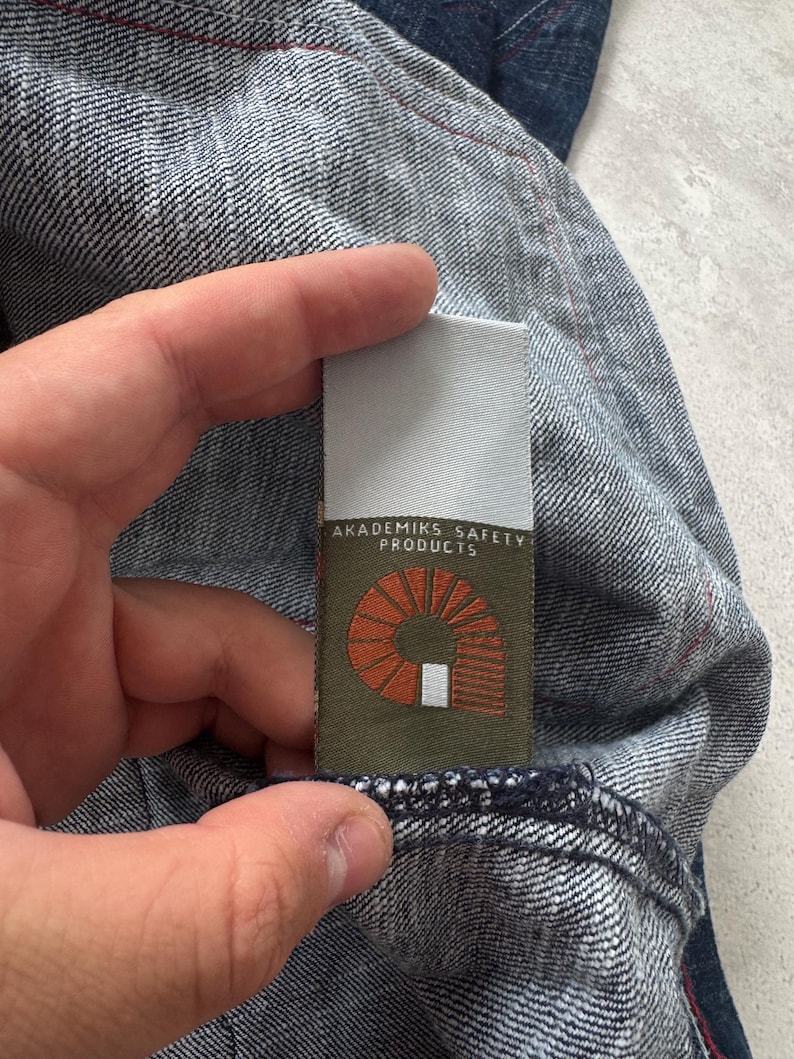 Może przedstawiać: Zbliżenie na tkaninę dżinsową z metką z napisem "AKADEMIKS SAFETY PRODUCTS". Etykieta ma pomarańczowy wz&oacute;r i biały tekst na zielonym tle. Dżins jest w średnim odcieniu z czerwonymi szwami.