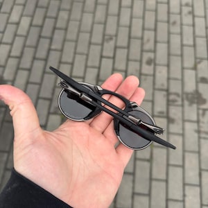 May include: Black sunglasses with round lenses and a silver frame detail. The sunglasses are folded and held in a hand. The background is a brick walkway.