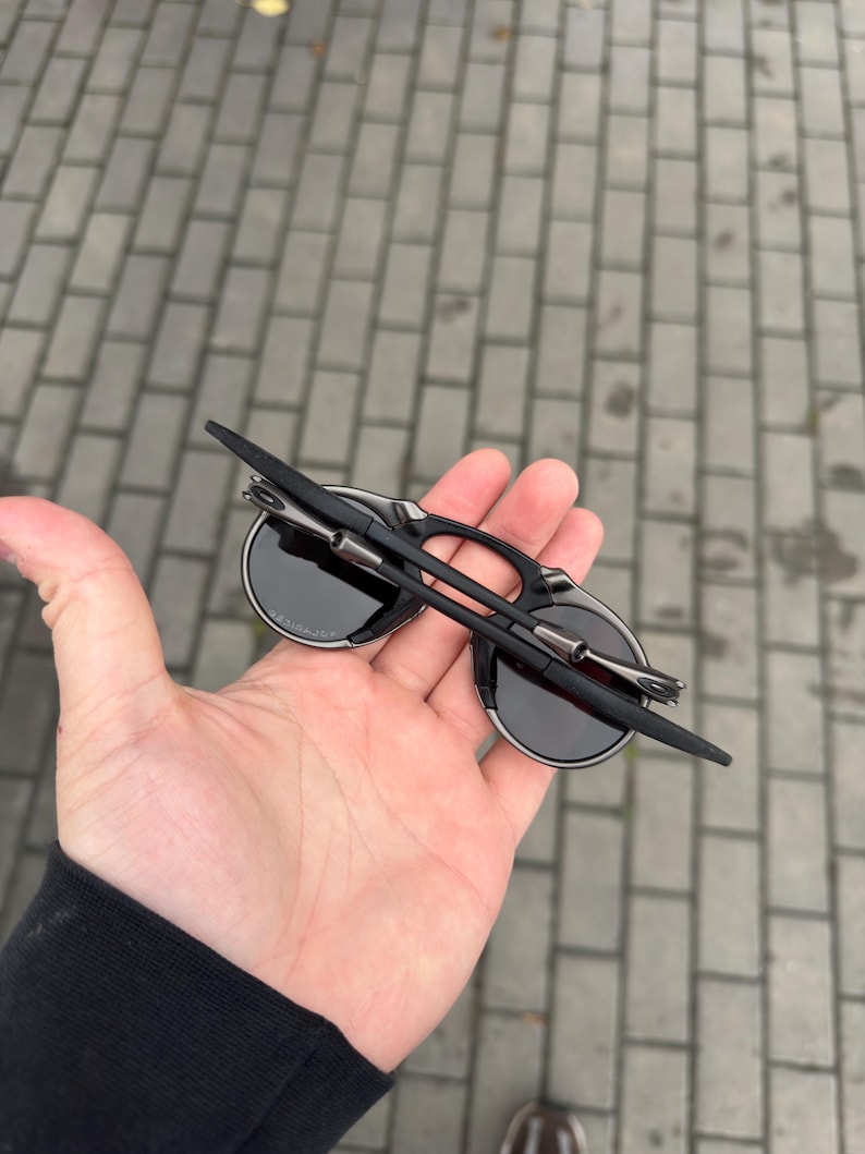 May include: Black sunglasses with round lenses and a silver frame detail. The sunglasses are folded and held in a hand. The background is a brick walkway.