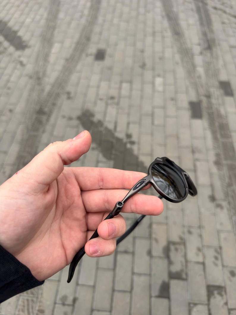 May include: Black rimless sunglasses with dark lenses. The sunglasses have a sleek, modern design with a thin, metallic frame and curved temples. The lenses reflect the surrounding environment.