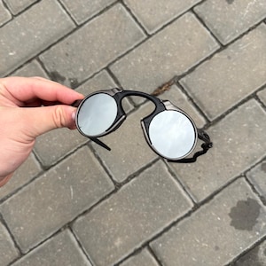 May include: Black and silver round sunglasses with a unique frame design. The lenses are reflective, and the frame is black with silver accents. The sunglasses are held against a gray brick background.