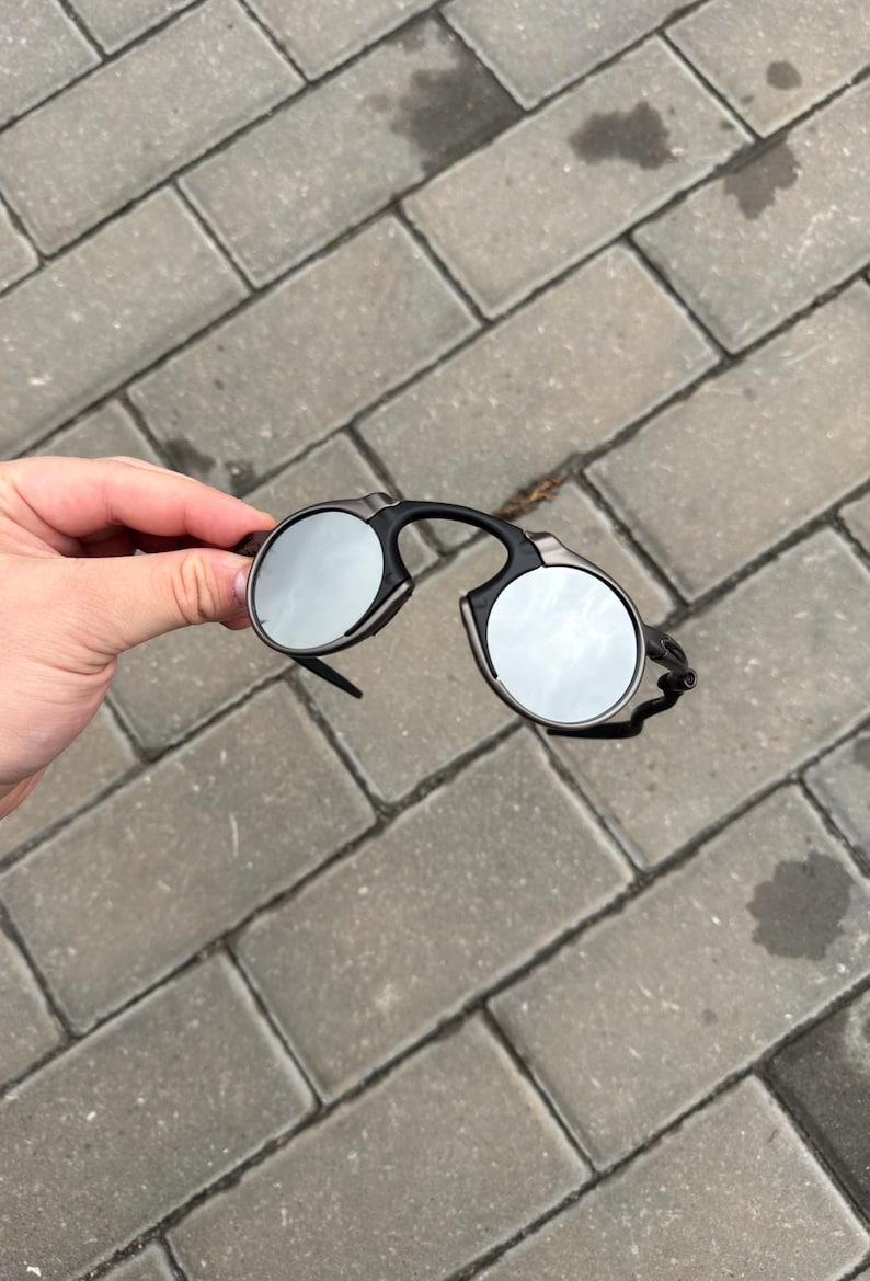 May include: Black and silver round sunglasses with a unique frame design. The lenses are reflective, and the frame is black with silver accents. The sunglasses are held against a gray brick background.