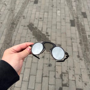 May include: Black rimless sunglasses with round, mirrored lenses. The sunglasses have a bridge and arms that connect to the lenses. The lenses reflect the surrounding environment.