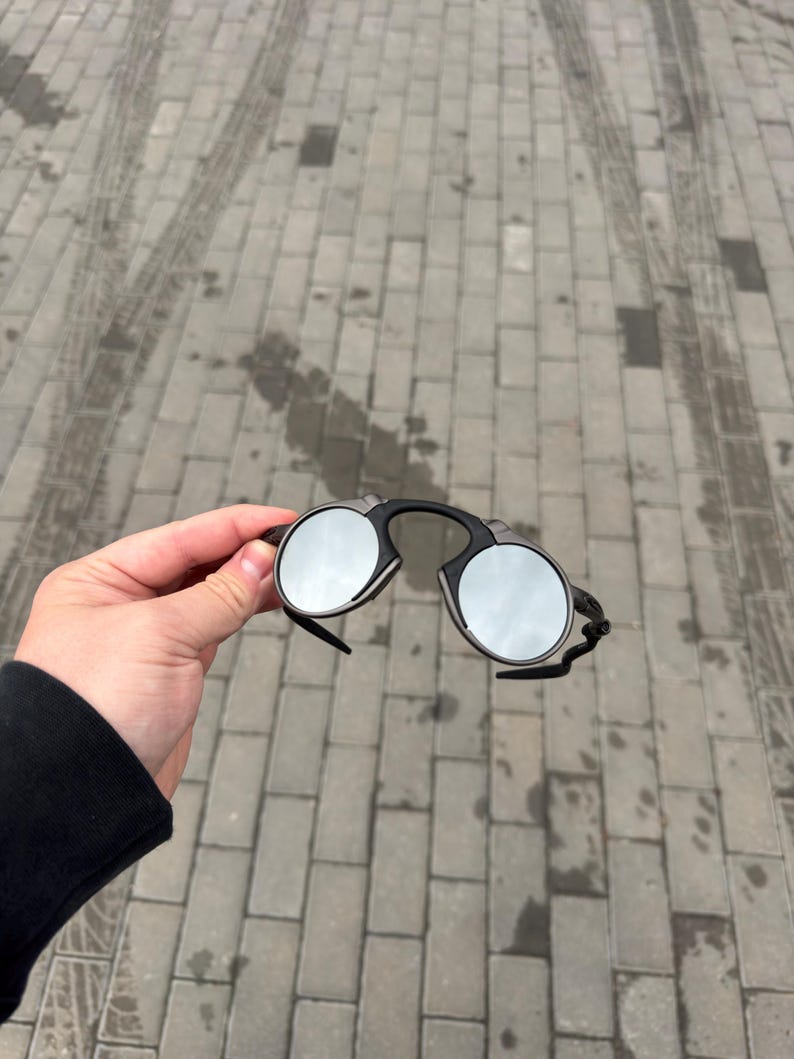 May include: Black rimless sunglasses with round, mirrored lenses. The sunglasses have a bridge and arms that connect to the lenses. The lenses reflect the surrounding environment.