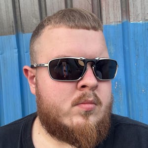 Puede incluir: Gafas de sol rectangulares con montura plateada y lentes oscuras. Las gafas tienen un puente y patillas negros. La persona de la imagen lleva una camiseta negra y tiene barba castaño rojiza.