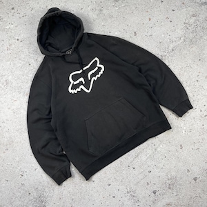 May include: A black long-sleeve hooded sweatshirt featuring a large white Fox Racing logo printed on the center chest. The garment includes a front kangaroo pocket, a drawstring hood with black cords, and ribbed cuffs at the sleeves and hem.